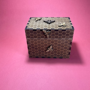May include: A wooden box with a honeycomb pattern and bee decorations. The box is made of dark wood and has a light brown honeycomb pattern. There are five bee decorations on the box, all of which are made of wood and have a light brown color.