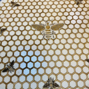May include: A round wooden serving board with a laser-etched design of a honeycomb pattern and several bees. The bees and honeycomb are a dark brown color against the light brown wood. The board is approximately 12 inches in diameter.