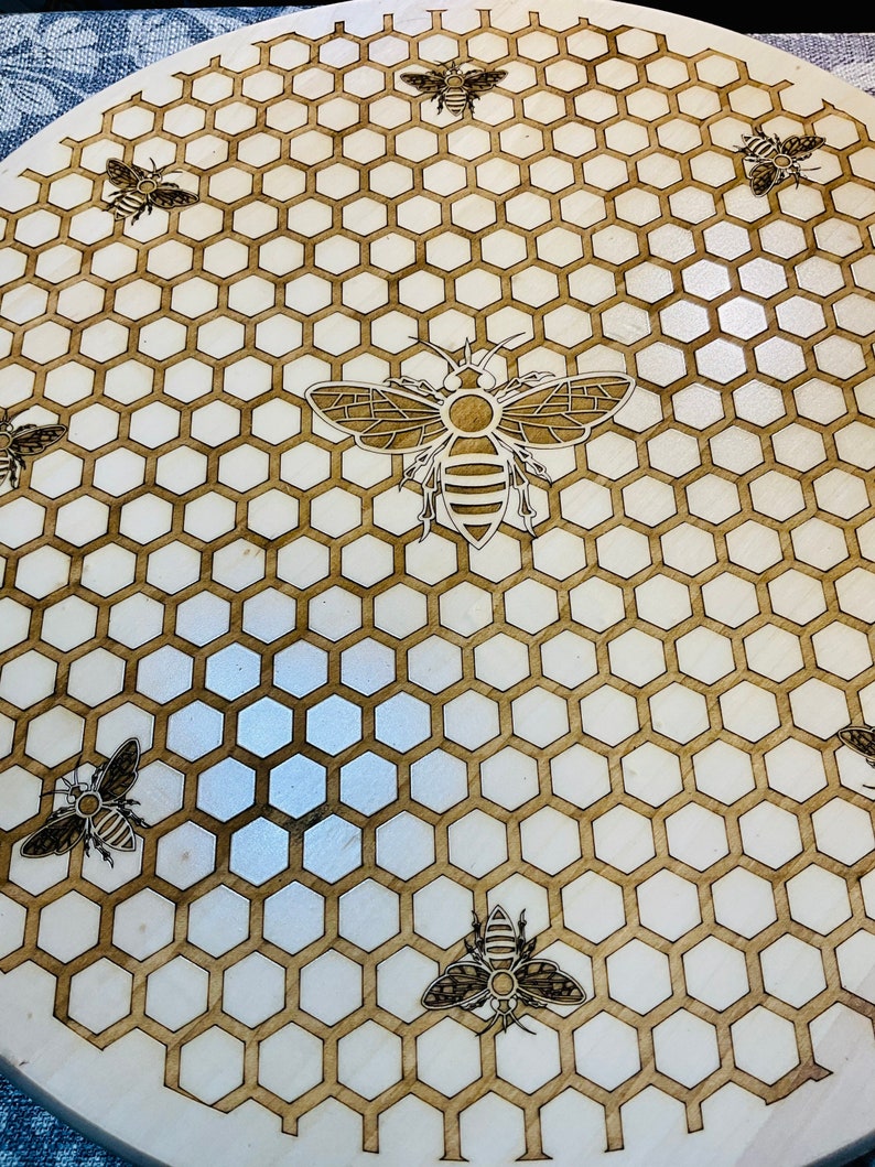 May include: A round wooden serving board with a laser-etched design of a honeycomb pattern and several bees. The bees and honeycomb are a dark brown color against the light brown wood. The board is approximately 12 inches in diameter.