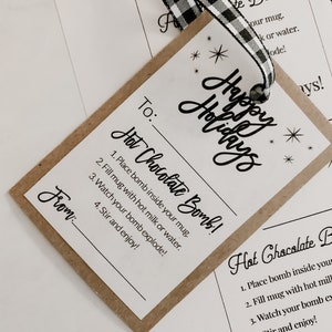 Puede incluir: Una etiqueta de regalo con las palabras "Felices Fiestas" e instrucciones para una bomba de chocolate caliente. La etiqueta es marr&oacute;n con texto blanco y una cinta a cuadros en blanco y negro.