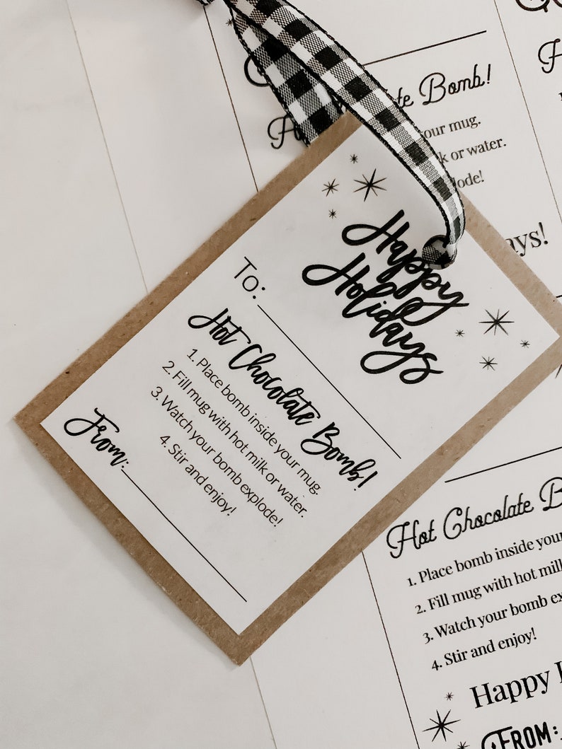 Puede incluir: Una etiqueta de regalo con las palabras "Felices Fiestas" e instrucciones para una bomba de chocolate caliente. La etiqueta es marr&oacute;n con texto blanco y una cinta a cuadros en blanco y negro.