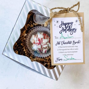 Puede incluir: Una caja de regalo festiva con una bomba de chocolate caliente. La bomba es blanca con rayas y trozos rojos, dentro de una taza transparente. La caja es de rayas plateadas y blancas. Una etiqueta dice "Happy Holidays" y "Hot Chocolate Bomb!"