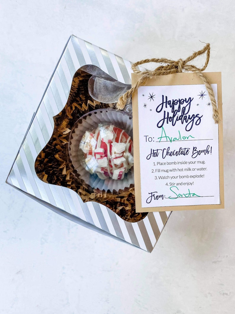 Puede incluir: Una caja de regalo festiva con una bomba de chocolate caliente. La bomba es blanca con rayas y trozos rojos, dentro de una taza transparente. La caja es de rayas plateadas y blancas. Una etiqueta dice "Happy Holidays" y "Hot Chocolate Bomb!"