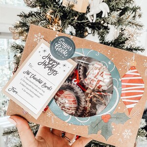 Puede incluir: Una caja festiva de bombas de chocolate caliente, ideal para las fiestas. La caja presenta una ventana transparente que muestra los dulces, con un dise&ntilde;o decorativo de adornos y acebo. Se incluye una tarjeta con instrucciones y el texto "Happy Holidays".
