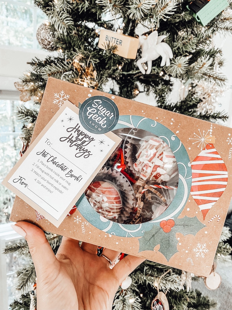 Puede incluir: Una caja festiva de bombas de chocolate caliente, ideal para las fiestas. La caja presenta una ventana transparente que muestra los dulces, con un dise&ntilde;o decorativo de adornos y acebo. Se incluye una tarjeta con instrucciones y el texto "Happy Holidays".