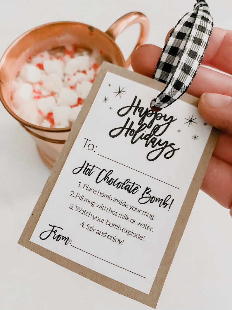 Puede incluir: Una taza de cobre llena de chocolate caliente y malvaviscos, junto a una etiqueta de regalo con las palabras "Happy Holidays" e instrucciones para una bomba de chocolate caliente. La etiqueta est&aacute; atada con una cinta a cuadros en blanco y negro.