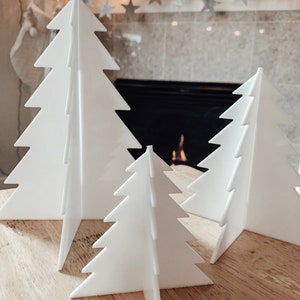 May include: A collection of three-dimensional white Christmas tree decorations. The trees are made of a flat material with a slotted design, allowing them to stand upright. The trees are placed on a wooden surface.