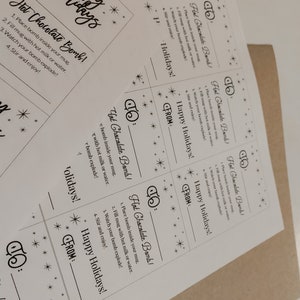 Puede incluir: Tarjetas de papel blanco con instrucciones para bombas de chocolate caliente. Las tarjetas tienen el texto "&iexcl;Felices Fiestas!" y "Hot Chocolate Bomb!" con decoraciones de estrellas.