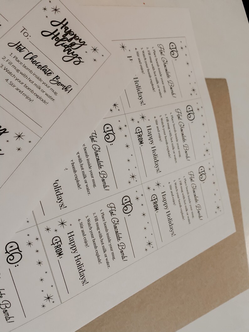 Puede incluir: Tarjetas de papel blanco con instrucciones para bombas de chocolate caliente. Las tarjetas tienen el texto "&iexcl;Felices Fiestas!" y "Hot Chocolate Bomb!" con decoraciones de estrellas.