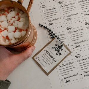 Puede incluir: Una taza de cobre llena de chocolate caliente y malvaviscos, junto a etiquetas impresas que dicen "Hot Chocolate Bomb!" y "Happy Holidays!". Las etiquetas est&aacute;n atadas con una cinta a cuadros en blanco y negro.