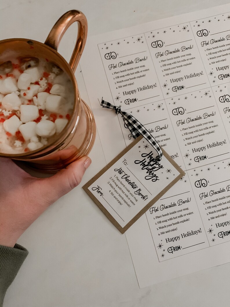 Puede incluir: Una taza de cobre llena de chocolate caliente y malvaviscos, junto a etiquetas impresas que dicen "Hot Chocolate Bomb!" y "Happy Holidays!". Las etiquetas est&aacute;n atadas con una cinta a cuadros en blanco y negro.
