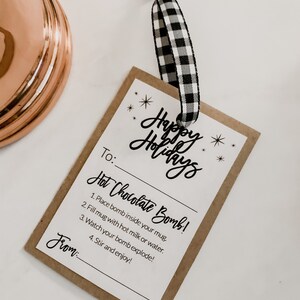 Puede incluir: Una etiqueta de regalo rectangular con las palabras "Happy Holidays" en escritura cursiva. La etiqueta incluye instrucciones para una bomba de chocolate caliente y est&aacute; sujeta a una cinta a cuadros en blanco y negro.