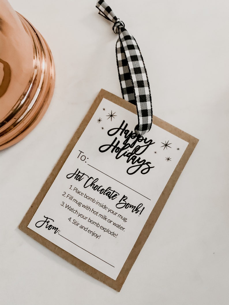 Puede incluir: Una etiqueta de regalo rectangular con las palabras "Happy Holidays" en escritura cursiva. La etiqueta incluye instrucciones para una bomba de chocolate caliente y est&aacute; sujeta a una cinta a cuadros en blanco y negro.
