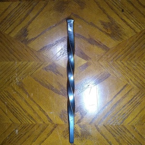 May include: A hand-forged, twisted metal bar with a square head and a flat end. The bar has a dark, metallic finish and a spiral design along its length. The object is likely a decorative or functional piece, possibly a handle or a decorative element.