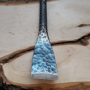 May include: A hand-forged metal chisel with a textured, hammered blade and a ridged handle. The blade has a blue-grey hue, and the handle is dark. The chisel rests on a wooden surface with a fur-like texture in the background.