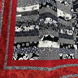 May include: A black and white patchwork quilt with a red border. The quilt is made of different fabrics with various patterns, including floral, geometric, and abstract designs.