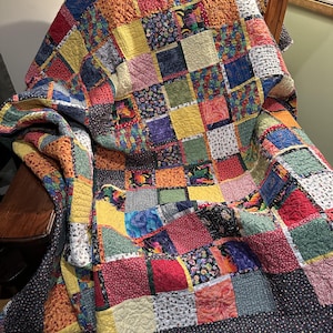 Puede incluir: Un edredón de patchwork hecho a mano, cubriendo una silla. El edredón presenta una vibrante mezcla de parches de tela cuadrados en varios colores y patrones, incluyendo texturas florales, geométricas y lisas. El borde del edredón es una tela oscura con pequeños puntos blancos.