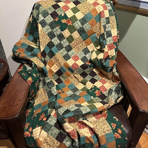 May include: A patchwork quilt with a variety of autumnal colours and patterns, including green, brown, orange, and yellow. The quilt is made of squares and features a leaf design. The quilt is draped over a wooden armchair.