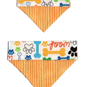 May include: Two reversible dog bandanas with a white background and a pattern of paw prints, bones, and the word "woof". The bandanas are orange on the reverse side with vertical stripes.