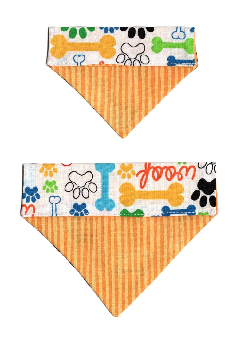 May include: Two reversible dog bandanas with a white background and a pattern of paw prints, bones, and the word "woof". The bandanas are orange on the reverse side with vertical stripes.