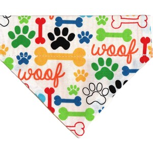 May include: White dog bandana with a colorful paw print and bone pattern. The bandana has the word "woof" printed on it twice.
