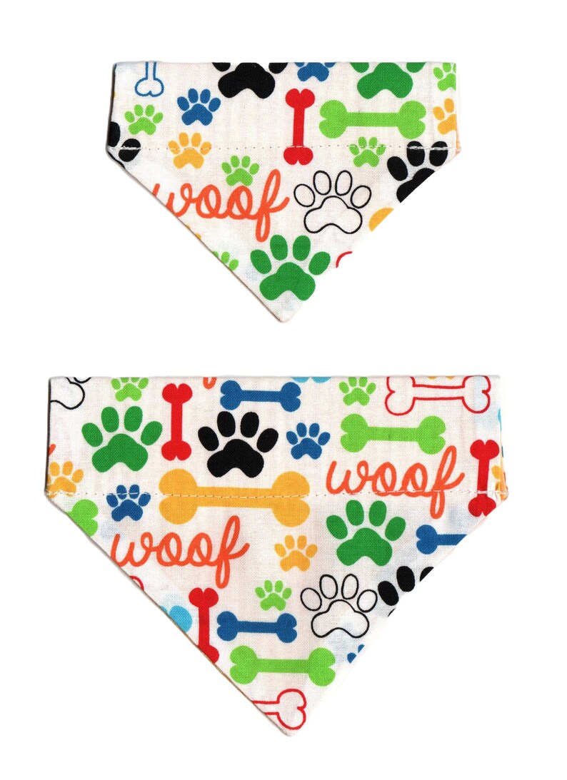May include: Two white dog bandanas with a colorful paw print and bone pattern. The bandanas have the word "woof" printed on them.