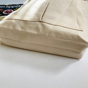 A beige canvas tote bag with 2 outer pockets and 1 inner pocket. The shoulder bag has a structured design with a flat bottom, spacious inside and long handles. The fabric appears to be a sturdy, natural canvas fabric.