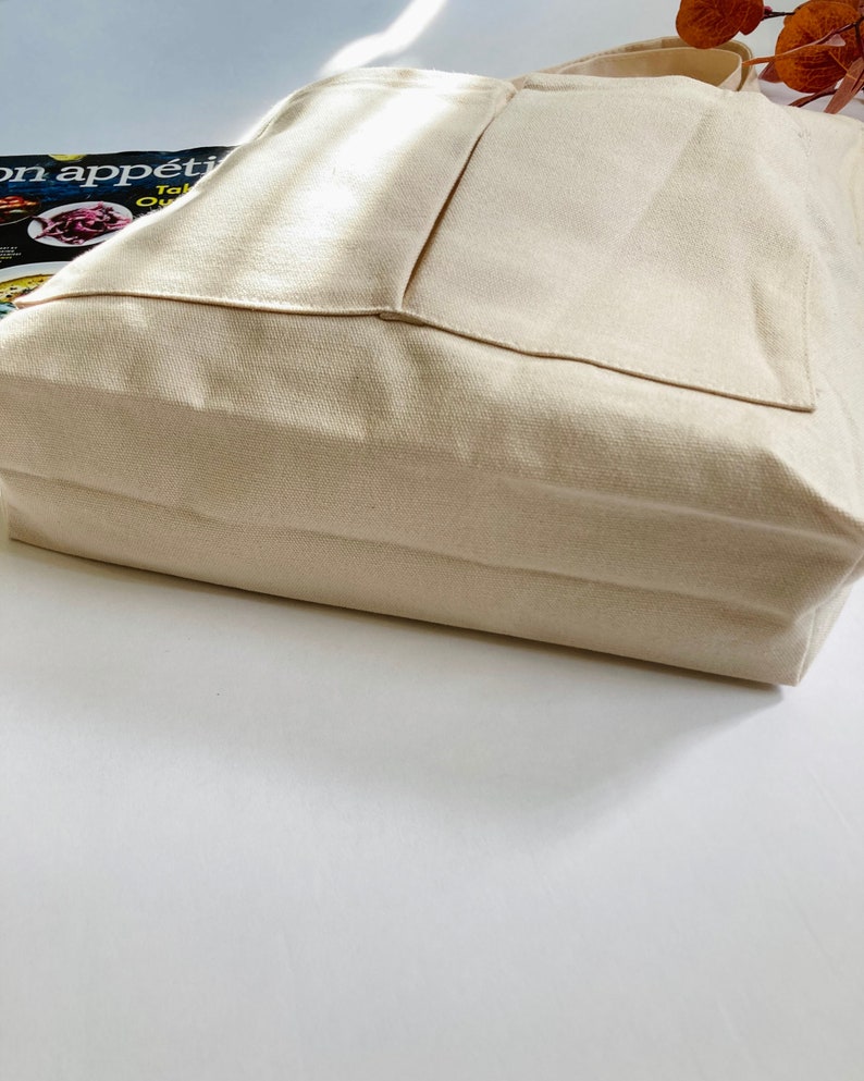 A beige canvas tote bag with 2 outer pockets and 1 inner pocket. The shoulder bag has a structured design with a flat bottom, spacious inside and long handles. The fabric appears to be a sturdy, natural canvas fabric.