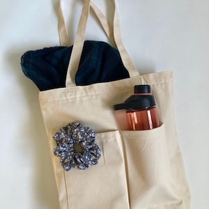 beige shoulder bag on white background with scarf inside and scrunchie outside. water bottle in one of the outer pockets of the bag