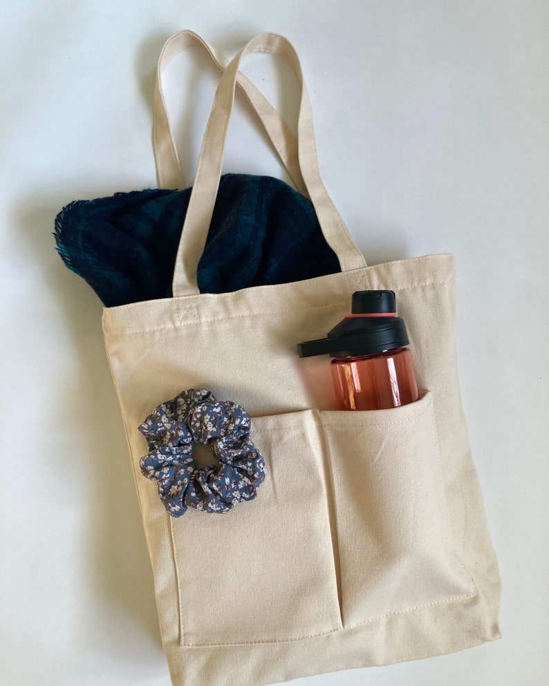beige shoulder bag on white background with scarf inside and scrunchie outside. water bottle in one of the outer pockets of the bag