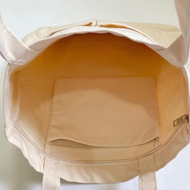 cream beige canvas bag showing shoulder straps, zipper, 2 front pockets, spacious inside with 1 inner pocket