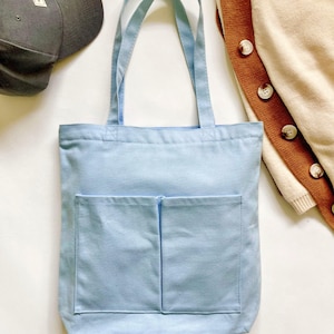 Light blue canvas tote bag with double front pockets and long shoulder straps, styled flat lay with gray cap and cream and brown cardigan, medium work and school carryall.