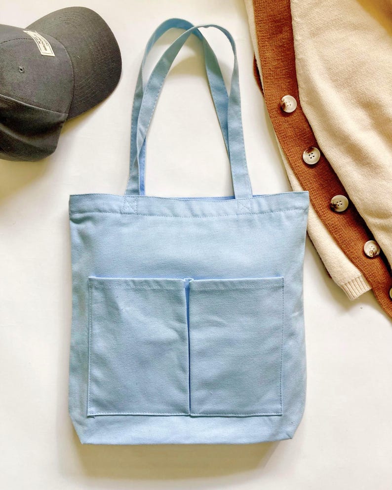 Light blue canvas tote bag with double front pockets and long shoulder straps, styled flat lay with gray cap and cream and brown cardigan, medium work and school carryall.