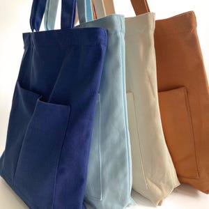 Four zippered canvas tote bags with front pockets in navy blue, light blue, cream, and burnt orange colors, standing side by side, medium 14x14 inch size with structured bottom and shoulder straps.