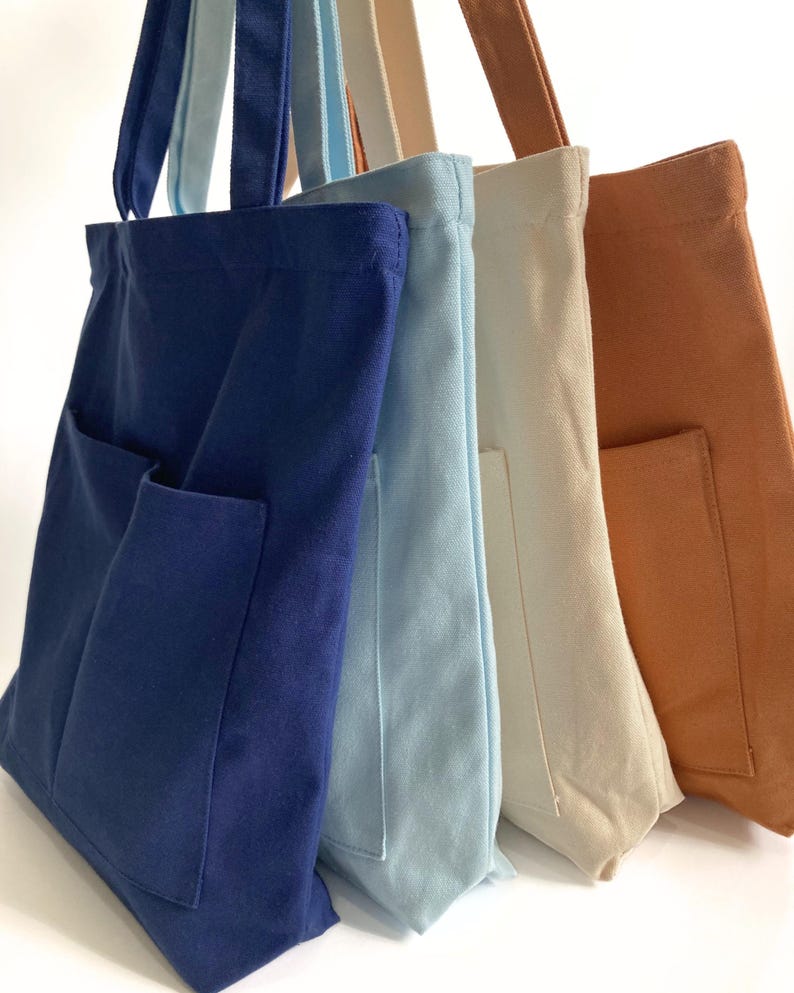Four zippered canvas tote bags with front pockets in navy blue, light blue, cream, and burnt orange colors, standing side by side, medium 14x14 inch size with structured bottom and shoulder straps.