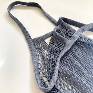 A gray reusable mesh shopping bag with a thick, woven strap. The bag is made of a fine, tightly woven mesh material.