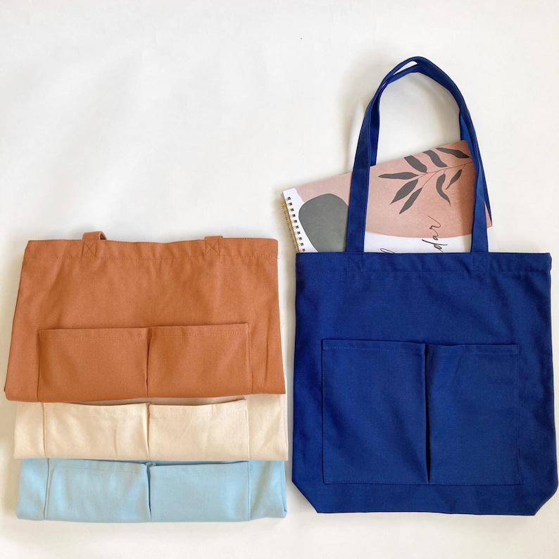 Navy blue zippered canvas tote bag with front pocket holding a notebook, shown with folded caramel brown, cream, and light blue color options, medium 14x14 inch work and school shoulder bag.