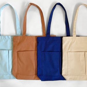 Four canvas tote bags in various colors: light blue, brown, navy blue, and beige. Each bag has a rectangular 2 front pocket and long sturdy handles. The bags are displayed against a white background.