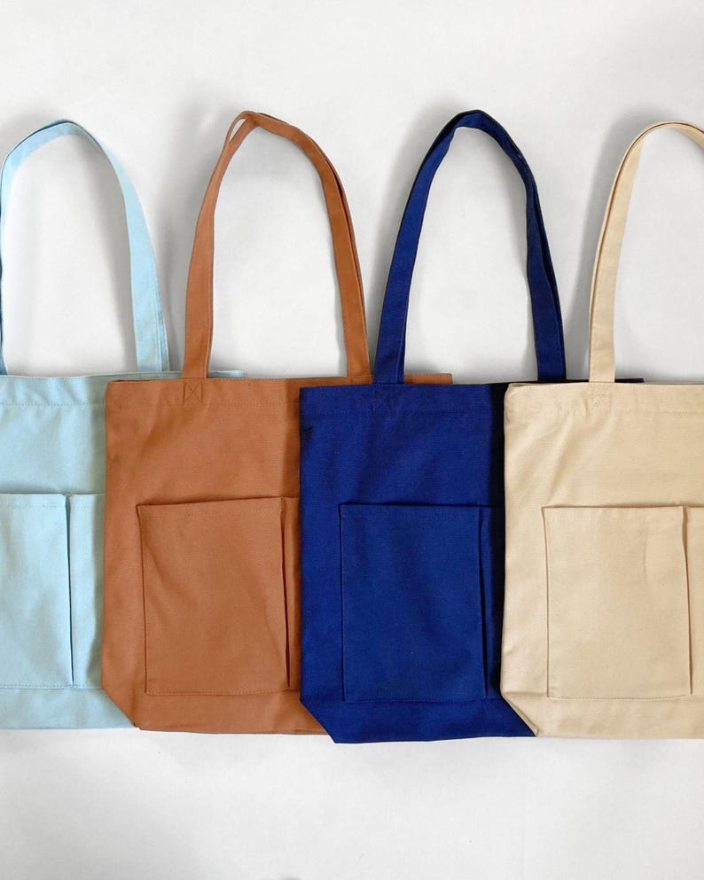 Four canvas tote bags in various colors: light blue, brown, navy blue, and beige. Each bag has a rectangular 2 front pocket and long sturdy handles. The bags are displayed against a white background.