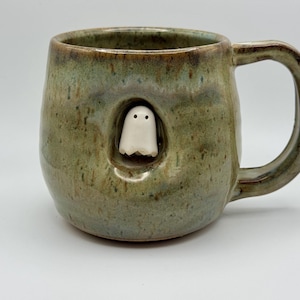May include: A handmade ceramic mug with a mottled green and brown glaze featuring a small, white, sculpted ghost figure peeking out from a recessed circular opening on the front side, complete with a sturdy handle on the right side.