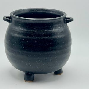 May include: A round, black ceramic pot with a speckled finish, featuring a wide opening, two small side handles, and three short, sturdy legs. The surface has a subtle horizontal ribbed texture throughout the body of the vessel.