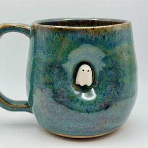 May include: A ceramic mug with a glossy, mottled teal and blue glaze featuring a small, sculpted white ghost figure emerging from a circular indentation on the side. The mug has a curved handle and a smooth, rounded body shape.
