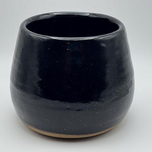 May include: A rounded ceramic cup with a glossy black glaze featuring subtle white speckles throughout. The vessel has a wide, open top and a smooth, curved body that tapers down to a natural, unglazed tan clay base at the bottom.