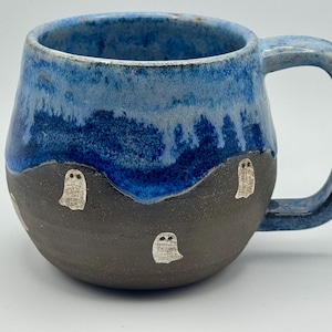 May include: A ceramic mug with a rounded shape, featuring a dark gray base decorated with small, white ghost illustrations. The upper portion of the mug is finished with a vibrant blue glaze that drips down over the dark gray surface.