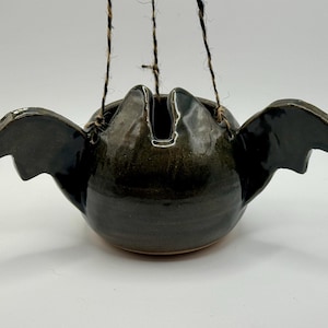 May include: A dark green, glossy ceramic hanging planter shaped like a bat with outstretched wings. The bowl features a rounded body and two pointed ears at the top rim. It is suspended by three thin, black and white twisted twine cords.