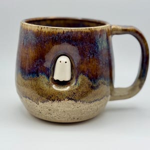 May include: A ceramic mug with a glossy, multi-toned glaze in shades of deep purple, brown, and tan. A small, white, three-dimensional ghost figure is embedded into the side of the mug, peeking out from a recessed circular indentation.