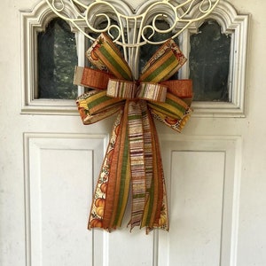May include: A large, decorative bow made of orange, green, and brown patterned ribbon is attached to a white door with a metal wreath hanger.