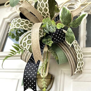 May include: A decorative bow featuring a mix of ribbons in solid olive green, natural tan burlap, black with white polka dots, and a white lattice pattern. The arrangement is accented with artificial green eucalyptus and fern foliage sprigs.