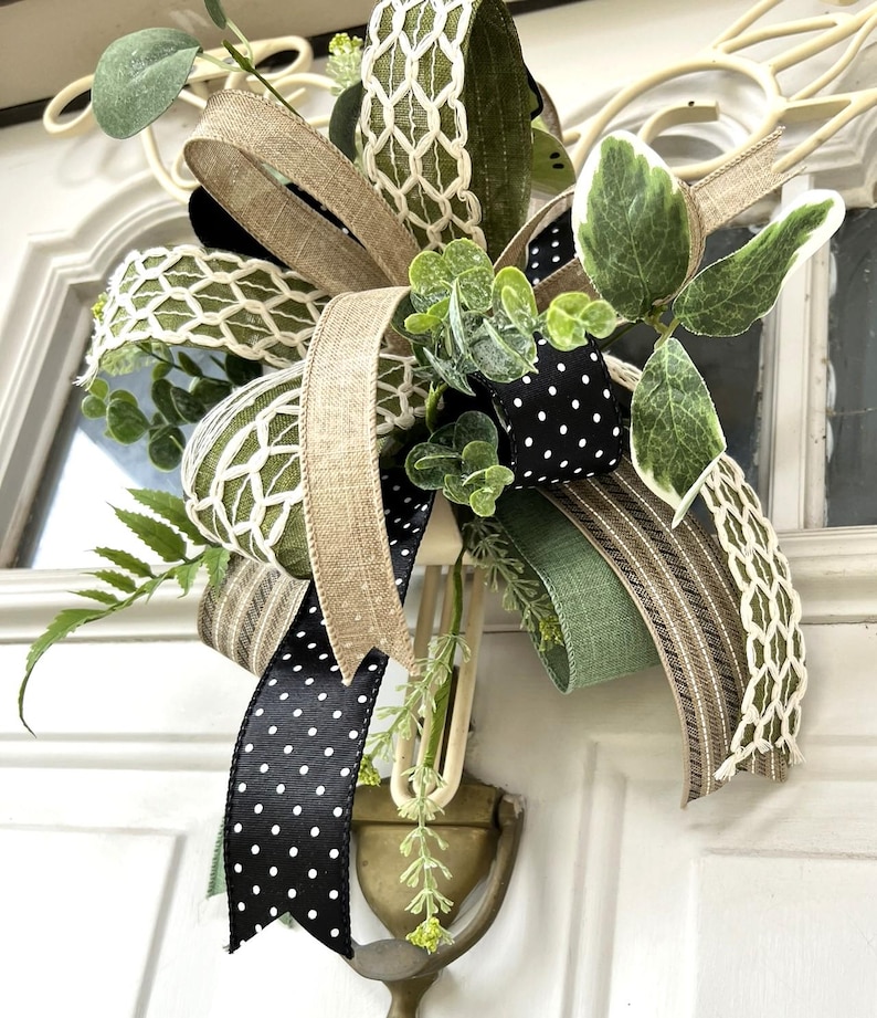May include: A decorative bow featuring a mix of ribbons in solid olive green, natural tan burlap, black with white polka dots, and a white lattice pattern. The arrangement is accented with artificial green eucalyptus and fern foliage sprigs.