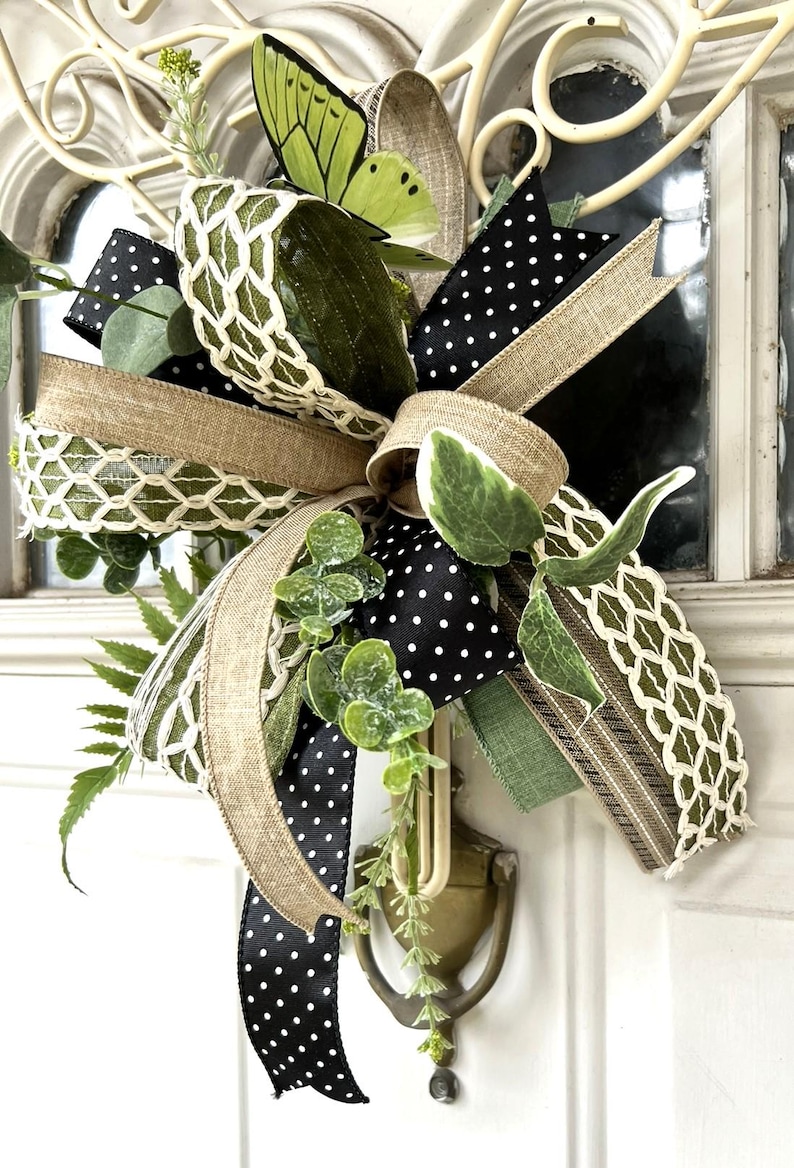 May include: A decorative bow featuring a mix of black and white polka dot ribbon, natural burlap ribbon, and green ribbon with a white lattice pattern. The arrangement includes artificial green eucalyptus leaves and a lime green butterfly accent.